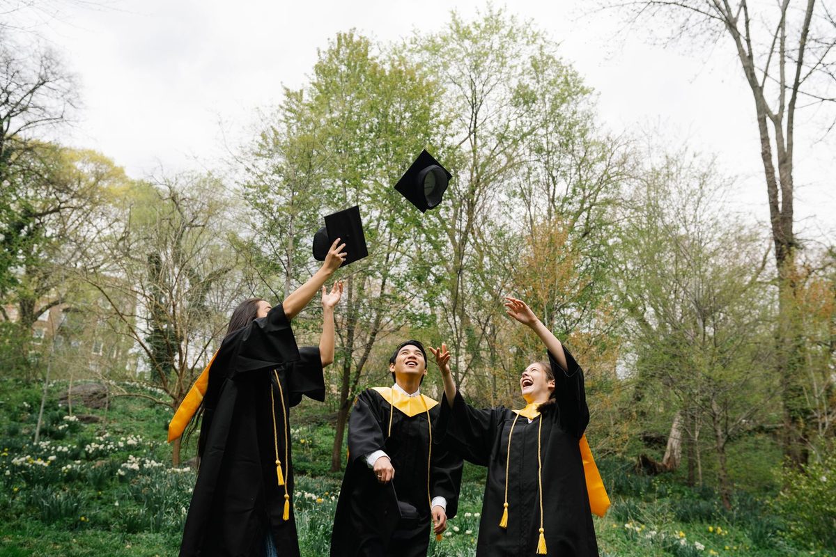 Three graduates in gowns tossing their caps in the air in a green park